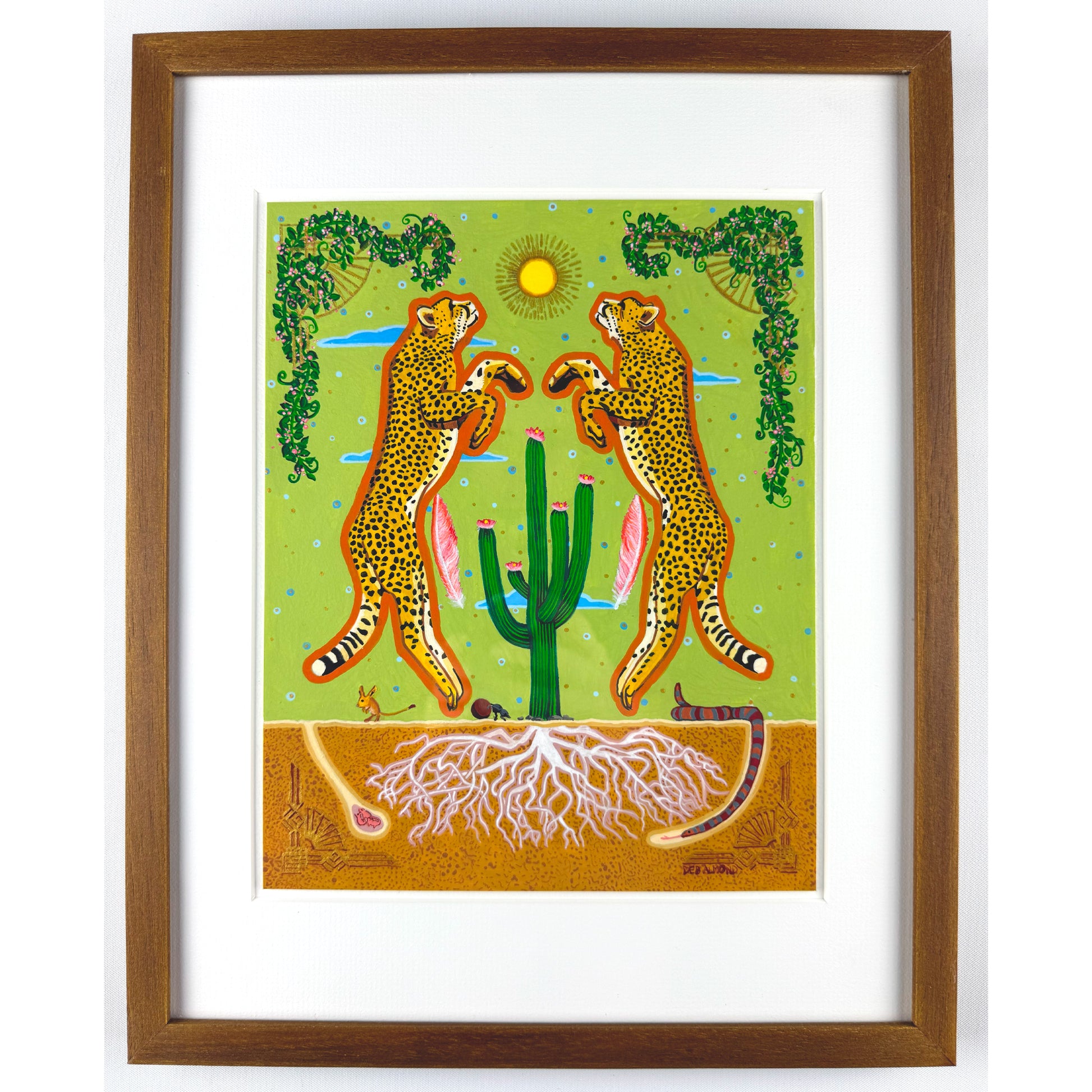 A colorful illustration shows two stylized cheetahs with tan fur and black spots standing upright, facing each other over a green cactus with pink blooms. The background features a light green sky with a yellow sun, blue clouds, and decorative green vines. Below the ground, white roots are visible in the brown soil, along with a desert mouse, a dung beetle, and a snake. The artwork is matted and framed in brown wood.