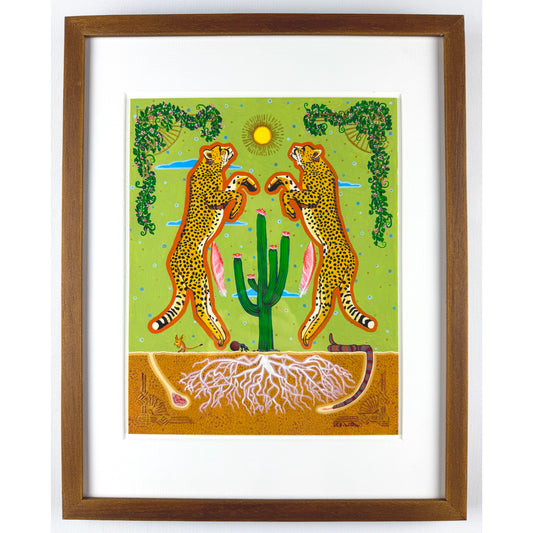 A colorful illustration shows two stylized cheetahs with tan fur and black spots standing upright, facing each other over a green cactus with pink blooms. The background features a light green sky with a yellow sun, blue clouds, and decorative green vines. Below the ground, white roots are visible in the brown soil, along with a desert mouse, a dung beetle, and a snake. The artwork is matted and framed in brown wood.