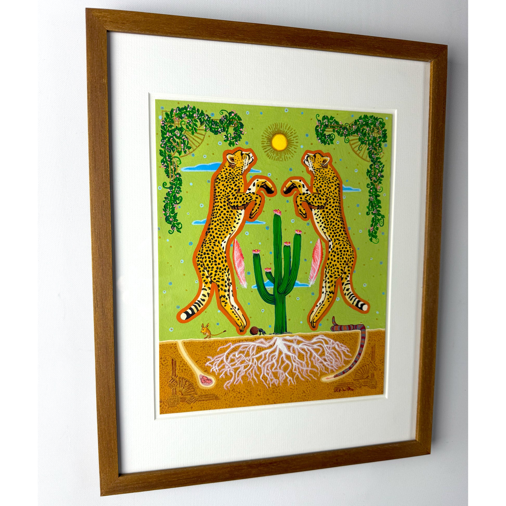 A slight angle of a colorful illustration shows two stylized cheetahs with tan fur and black spots standing upright, facing each other over a green cactus with pink blooms. The background features a light green sky with a yellow sun, blue clouds, and decorative green vines. Below the ground, white roots are visible in the brown soil, along with a desert mouse, a dung beetle, and a snake. The artwork is matted and framed in brown wood.
