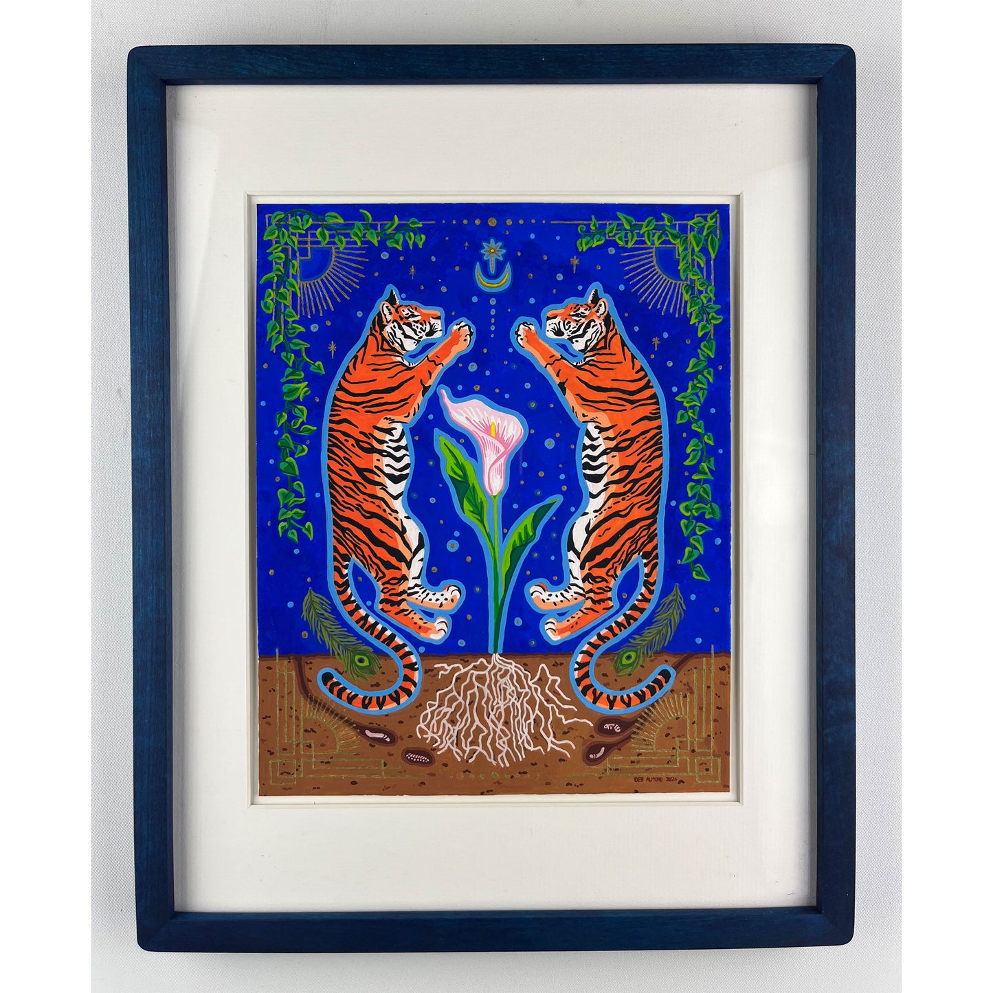 A vibrant illustration features two orange and black striped tigers standing upright and mirroring each other. Between them grows a pink calla lily with green leaves and exposed white roots in brown soil. The background is a deep blue night sky with a crescent moon, stars, and decorative green vines. The artwork is matted and framed in a dark blue frame.