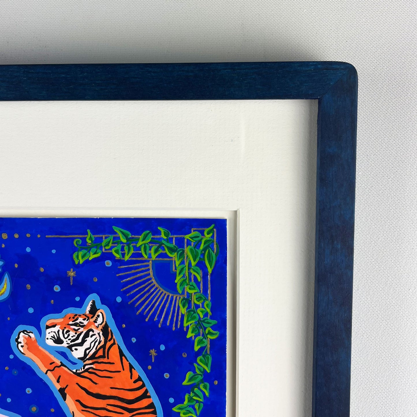 The top right corner of a vibrant illustration featuring one of two orange and black striped tigers standing upright. The background is a deep blue night sky and decorative green vines. The artwork is matted and framed in a dark blue frame.