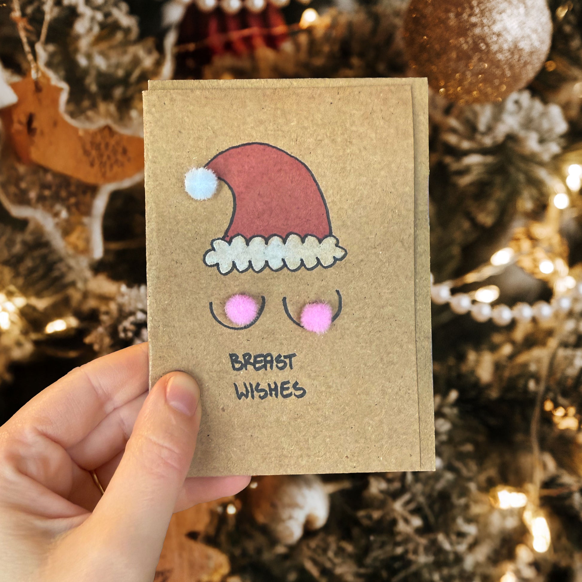 Hand holding a small brown card with a Santa hat design and 'Breast Wishes' text, against a festive background.