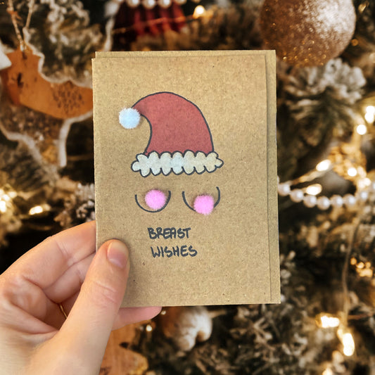 Hand holding a small brown card with a Santa hat design and 'Breast Wishes' text, against a festive background.