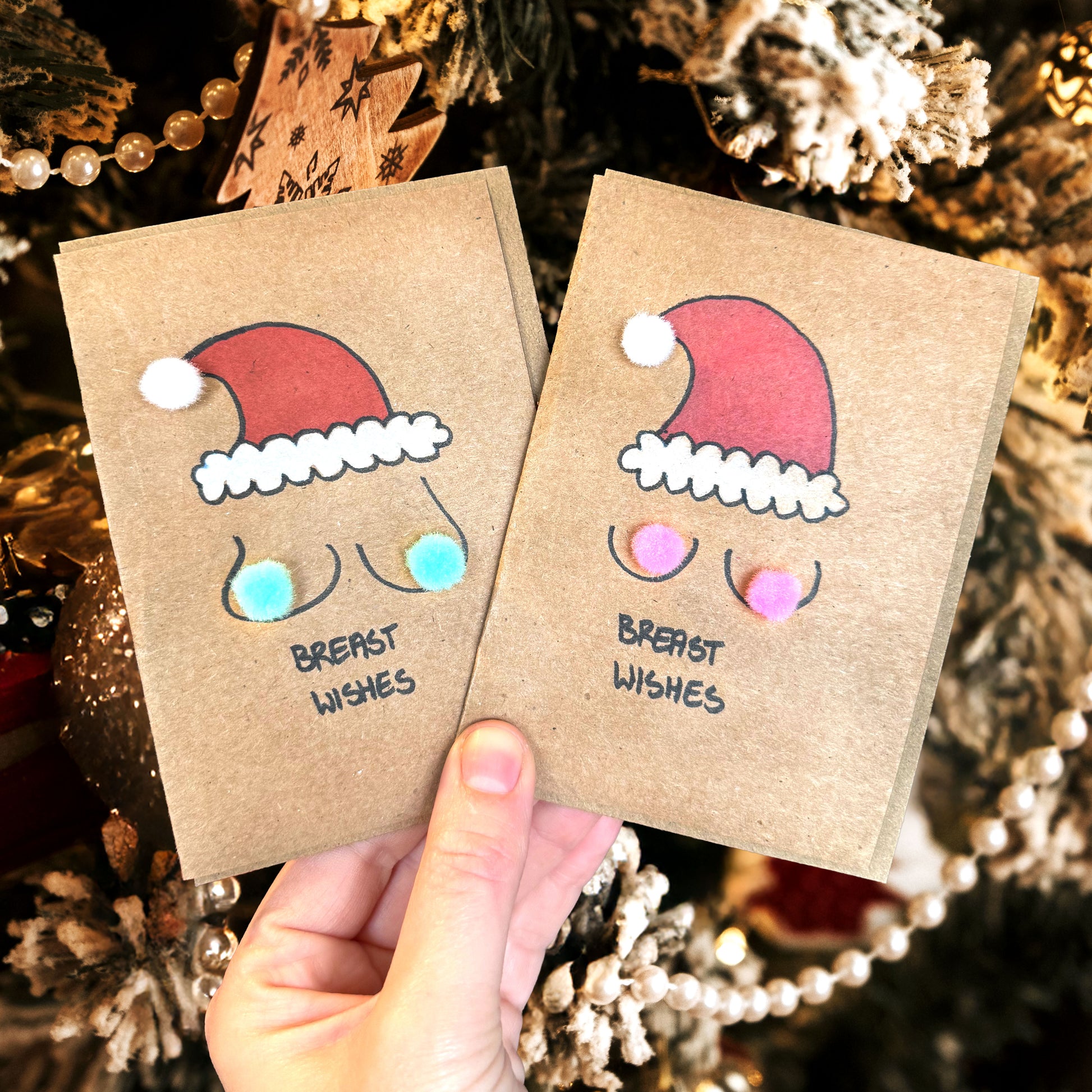 Two small brown cards with hand-drawn santa hats and 'Breast Wishes' text with pompoms, held in front of a decorated Christmas tree.