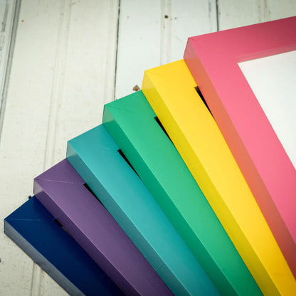 A close-up of a set of colorful picture frames fanned out diagonally, showing vibrant shades of blue, purple, teal, green, yellow, and pink against a white wood plank background.