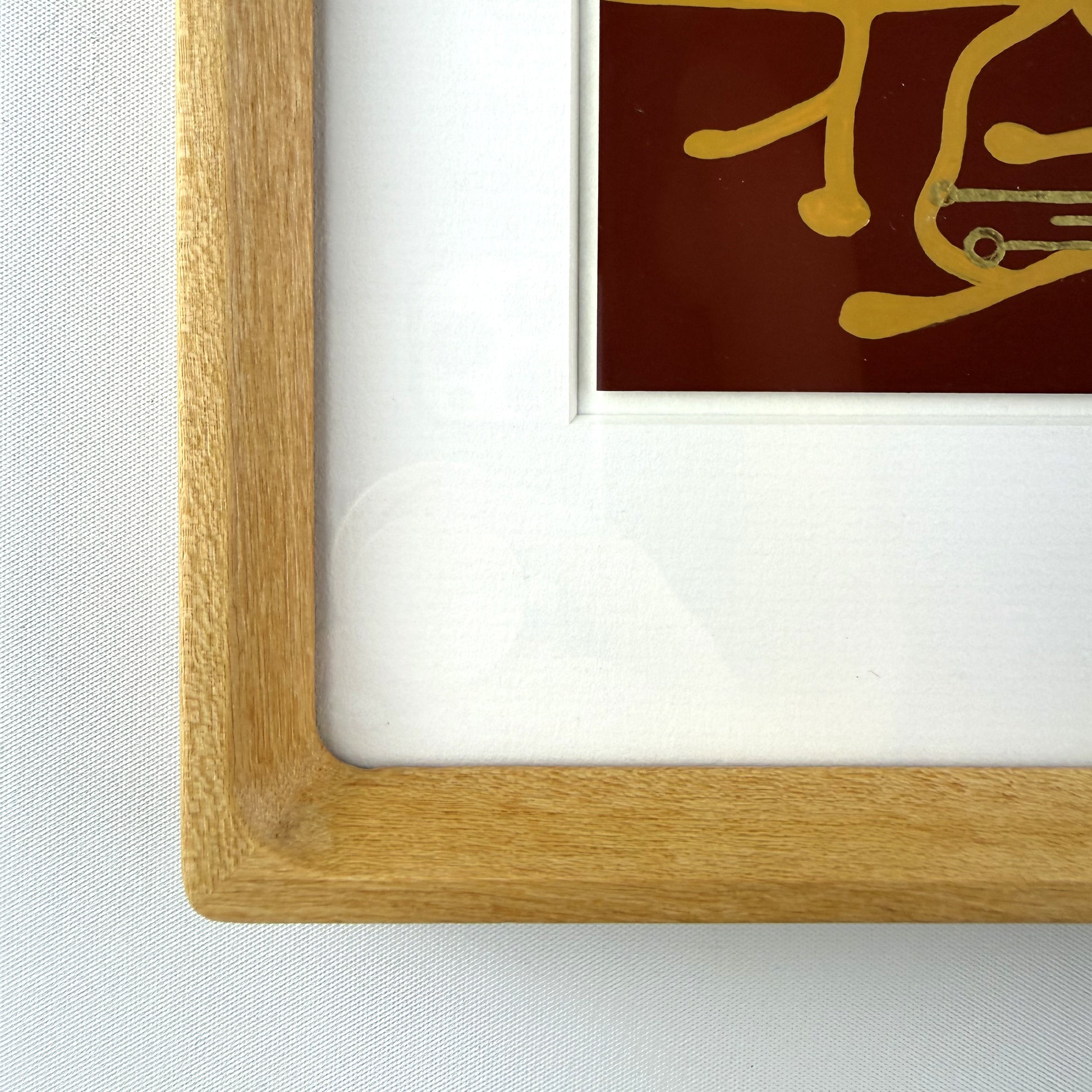 Close-up of a light oak wood picture frame with a white mat and a piece of wall art featuring gold details on a dark red background.