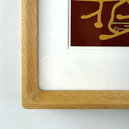 Close-up of a light oak wood picture frame with a white mat and a piece of wall art featuring gold details on a dark red background.