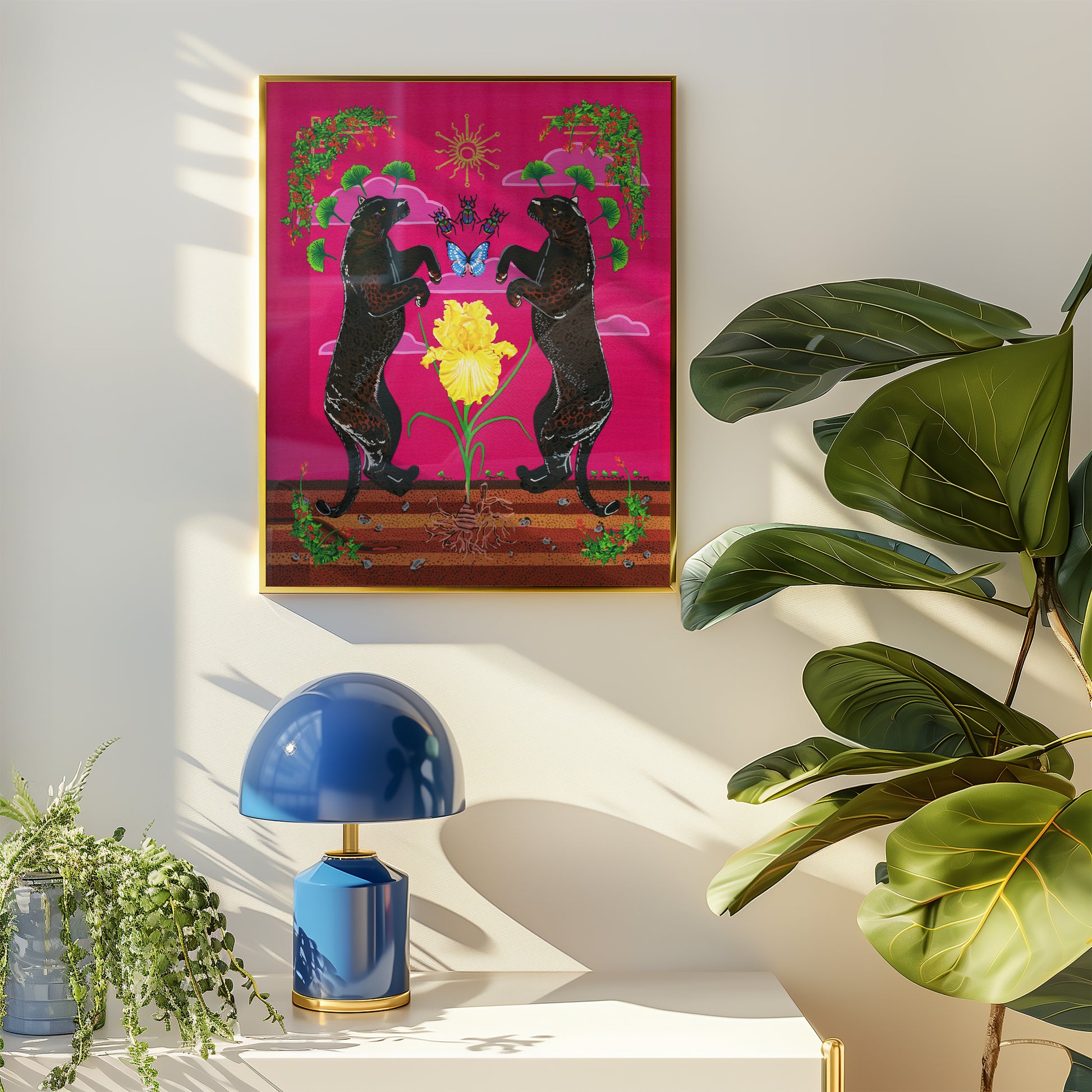 Framed giclée print in a thin gold frame hanging on a off-white wall, styled above a white side table with a blue dome lamp and lush green houseplants under natural sunlight.