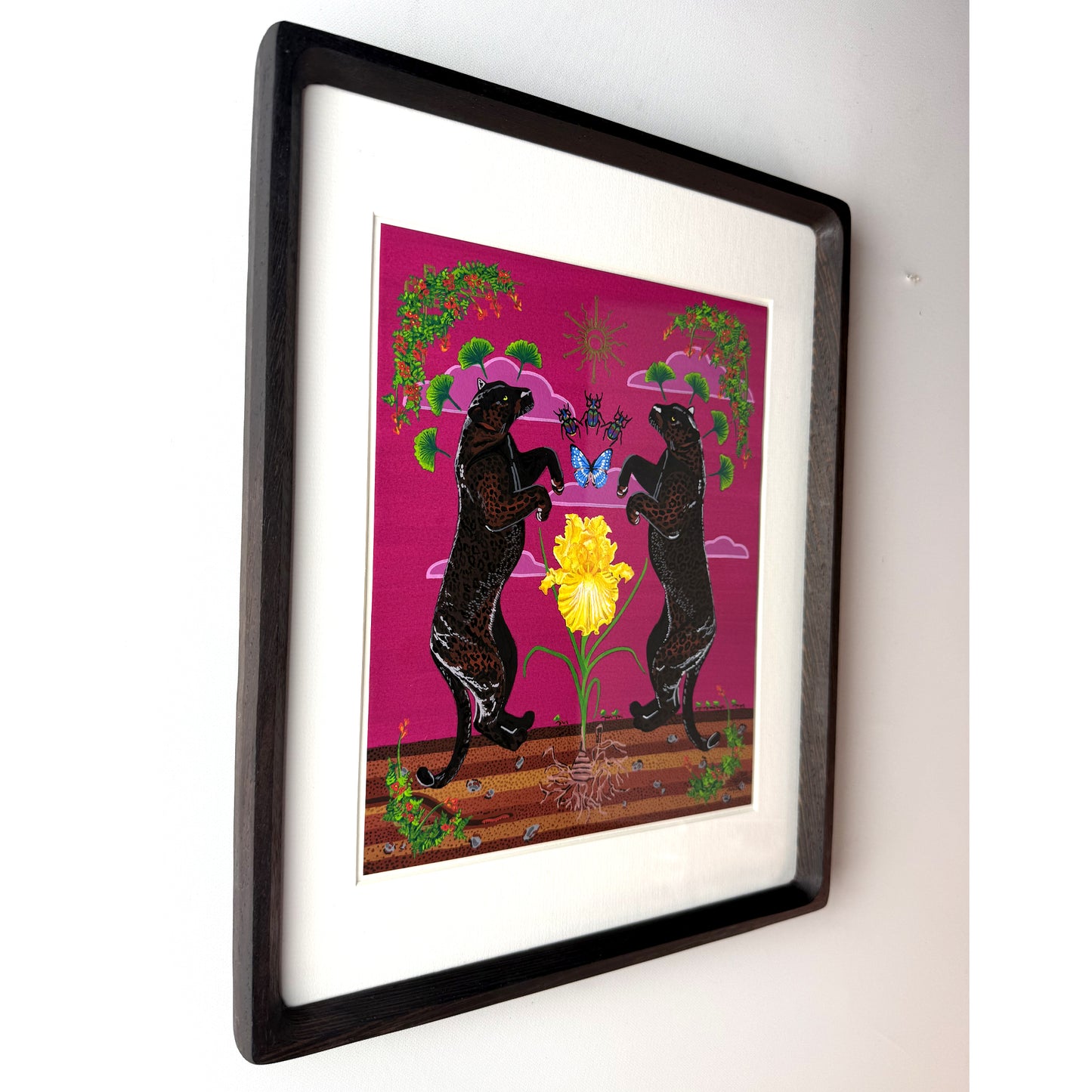 Framed art print featuring two black panthers standing on hind legs around a large yellow iris against a bright pink background, mounted in a dark wood frame with a white mat on a light wall.
