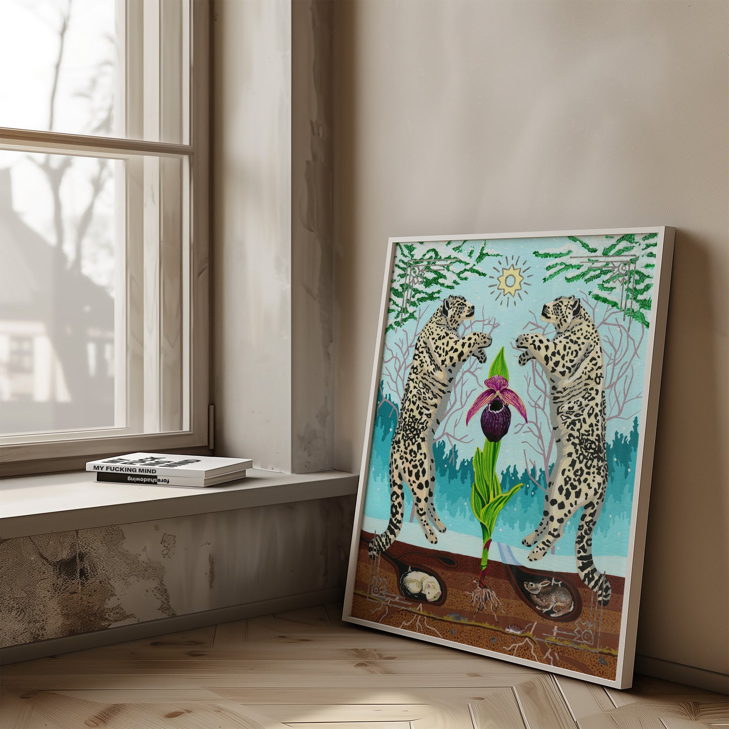 A framed surreal art print leaning against a beige wall on a light wood floor, next to a window. The artwork shows two leopards facing a large purple and pink orchid flower, with green leaves, a blue forest background, and a yellow sun.