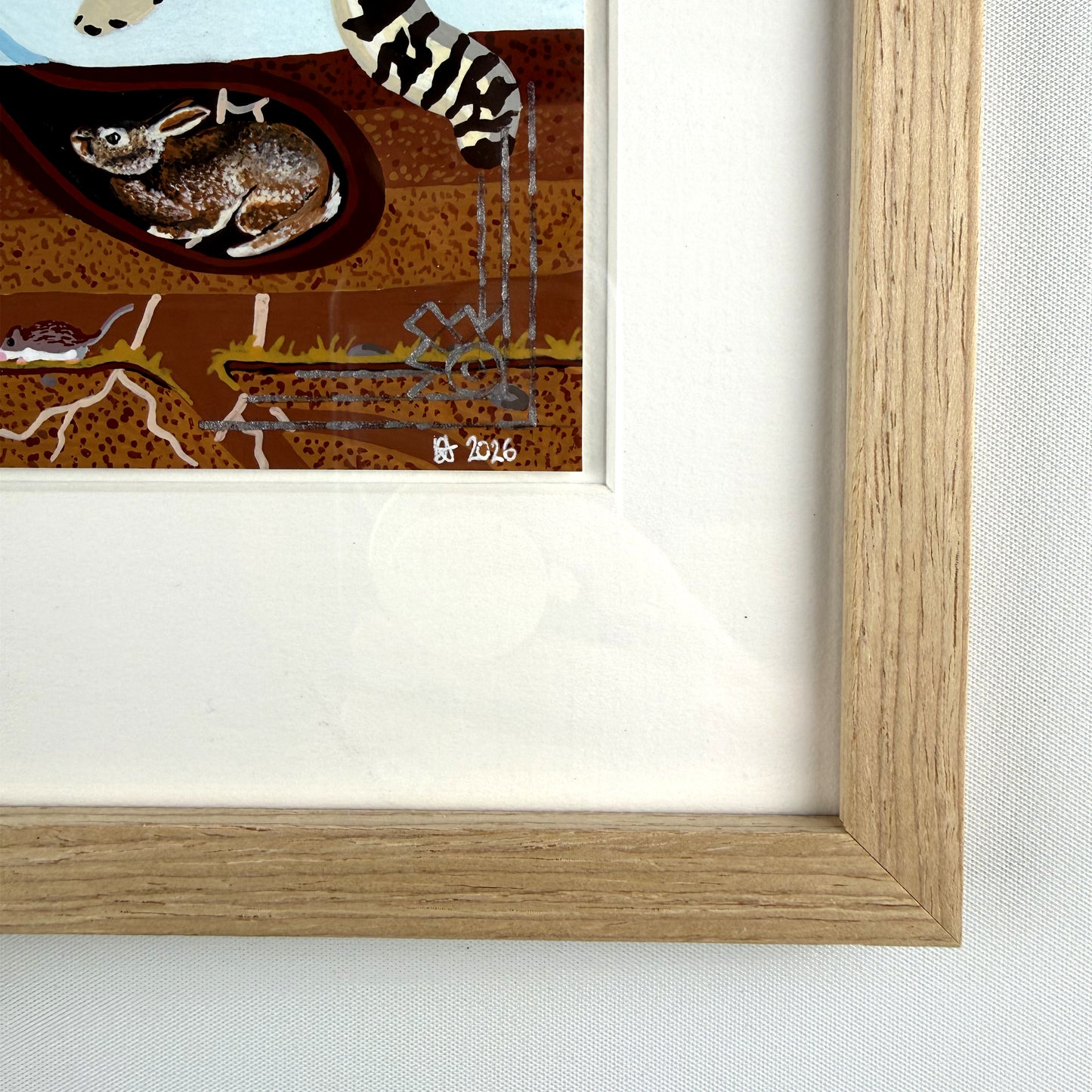 Close-up shot of the bottom-right corner of the light wood frame and white mat, highlighting the signature, date "2026", metallic detailing, and the underground burrow with a curled-up rodent.