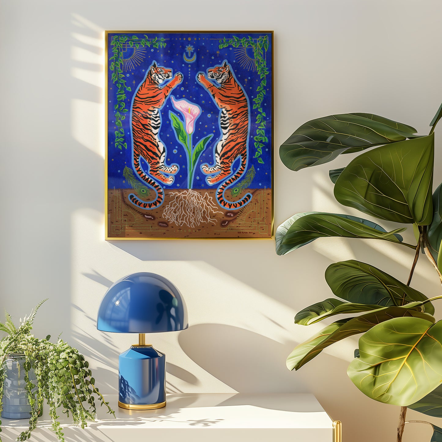 Framed giclée print in a thin gold frame mounted on a white wall above a console table, styled with a glossy blue table lamp and lush green houseplants in natural sunlight.