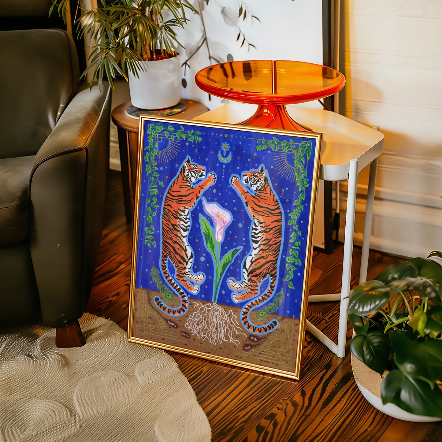 Framed giclée print resting on the floor in a gold frame next to an armchair and side table, set against dark hardwood floors and potted indoor plants.